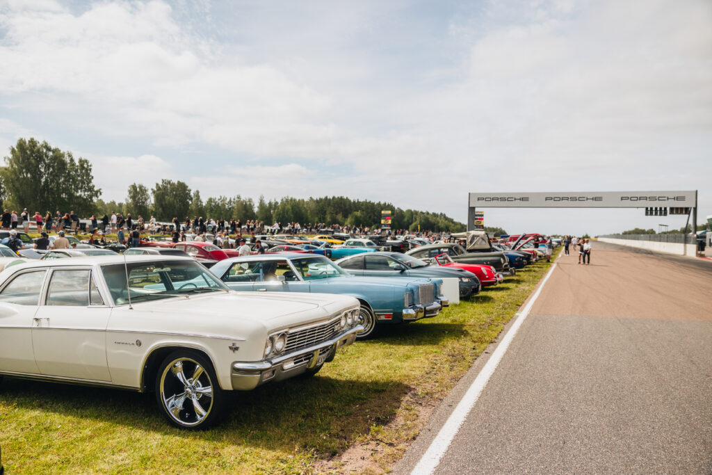 Klassiska bilar står parkerade i rader på gräset intill en racerbana, med en folkmassa i bakgrunden och en bro med Porsche-logotypen som sträcker sig över banan en delvis molnig dag.