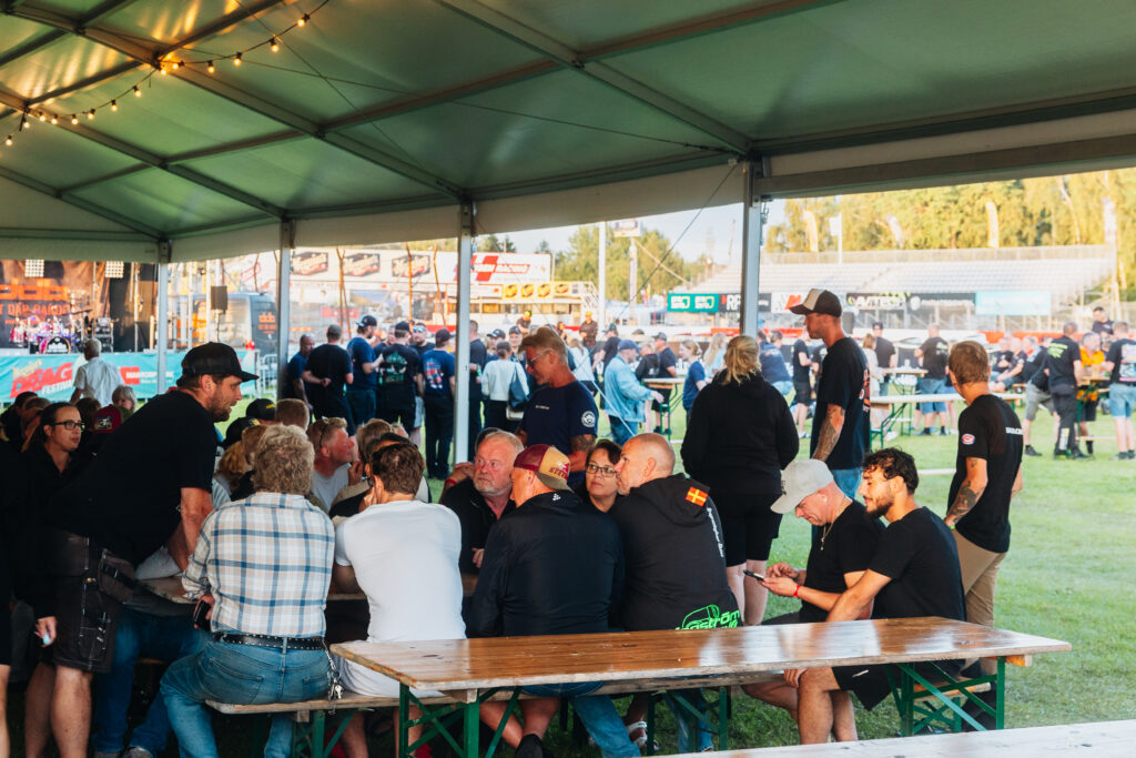 Människor sitter och står under ett stort tält vid ett utomhusevenemang och pratar och umgås vid träbord. I bakgrunden syns fler deltagare och evenemangsmontrar en solig dag.