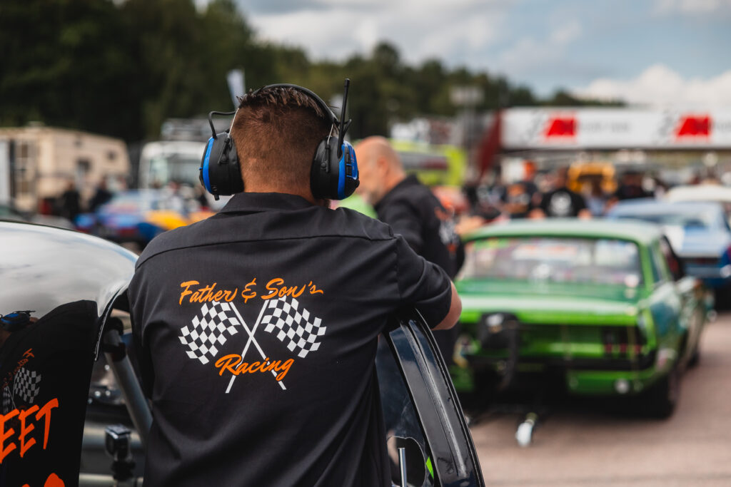 En man med hörlurar och en tröja från Father & Son’s Racing lutar sig mot en bil på en racerbana. Suddiga bilar och människor i bakgrunden ger en känsla av den livliga stämningen under en tävlingsdag.