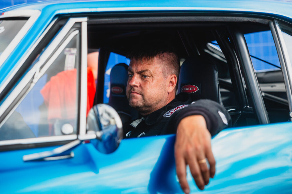 En man klädd i en racerdräkt sitter i förarsätet i en blå bil, med armen vilande mot dörren och med ett allvarligt uttryck i ansiktet.