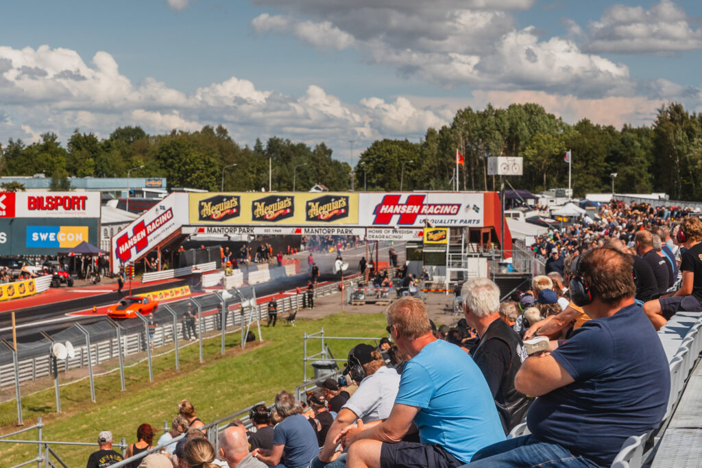 Åskådare tittar på ett dragrace på en racerbana en solig dag. Två bilar susar fram längs banan, medan publiken sitter på utomhusläktarna. Reklambanderoller kantar banan och i bakgrunden syns träd.