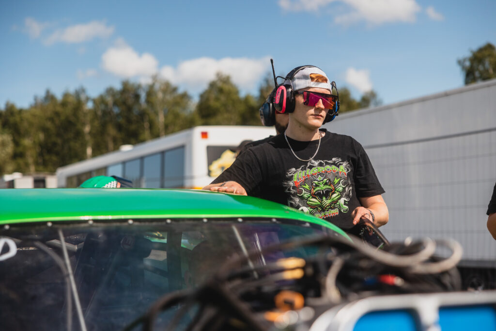 En person med solglasögon, hörlurar och en svart t-shirt står bredvid en grön racerbil utomhus en solig dag, med träd och husvagnar i bakgrunden.