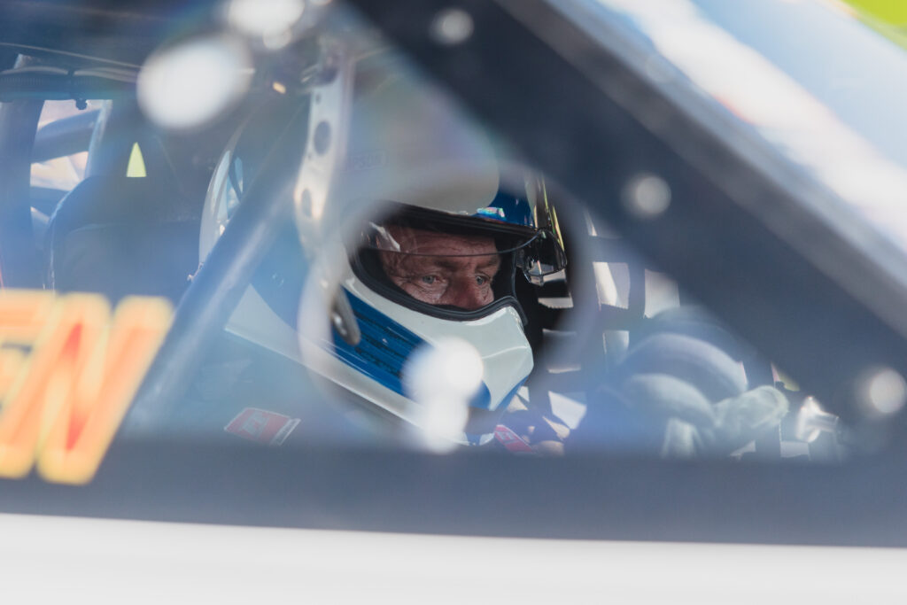 En racerförare med hjälm sitter i bilen och blickar rakt fram; bilden är tagen genom sidorutan.