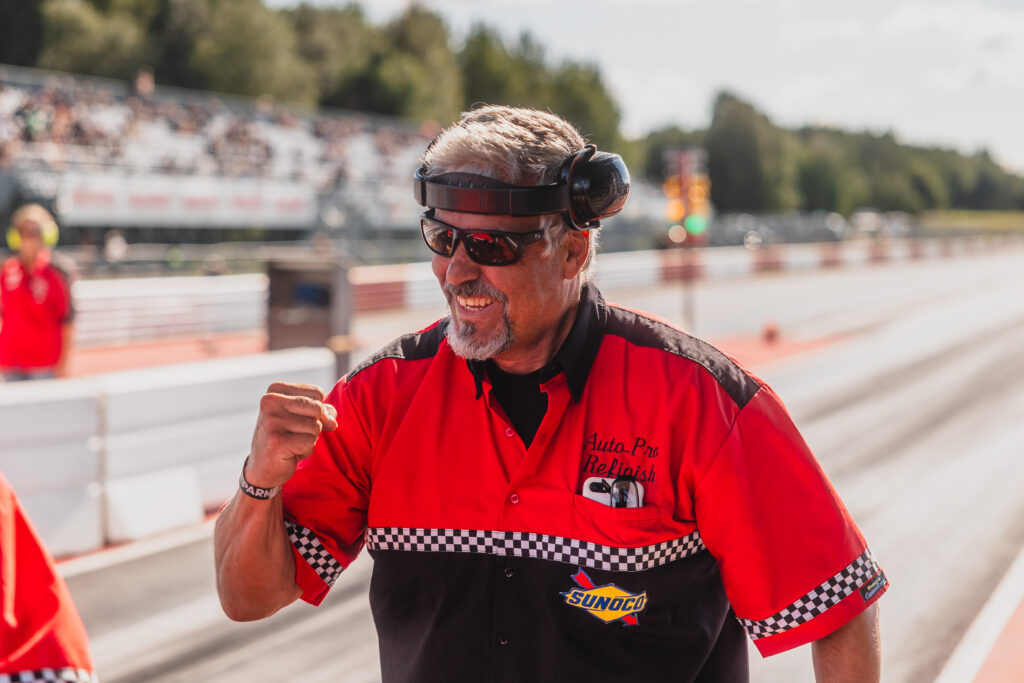 En man i en röd och svart racingtröja ler och lyfter näven i jubel på en racerbana. Han har hörlurar och solglasögon på sig, och i bakgrunden syns suddiga åskådare.