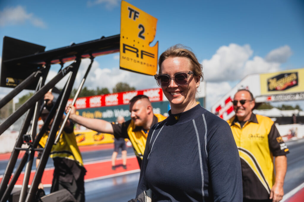En kvinna i racingdräkt och solglasögon ler på en racerbana, medan tre teammedlemmar i gula uniformer och racingutrustning syns i bakgrunden under en solig himmel.