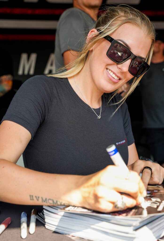 En kvinna med solglasögon och en svart tröja ler medan hon skriver autografer vid ett bord. Hon har ljust hår uppsatt och en synlig tatuering på underarmen.