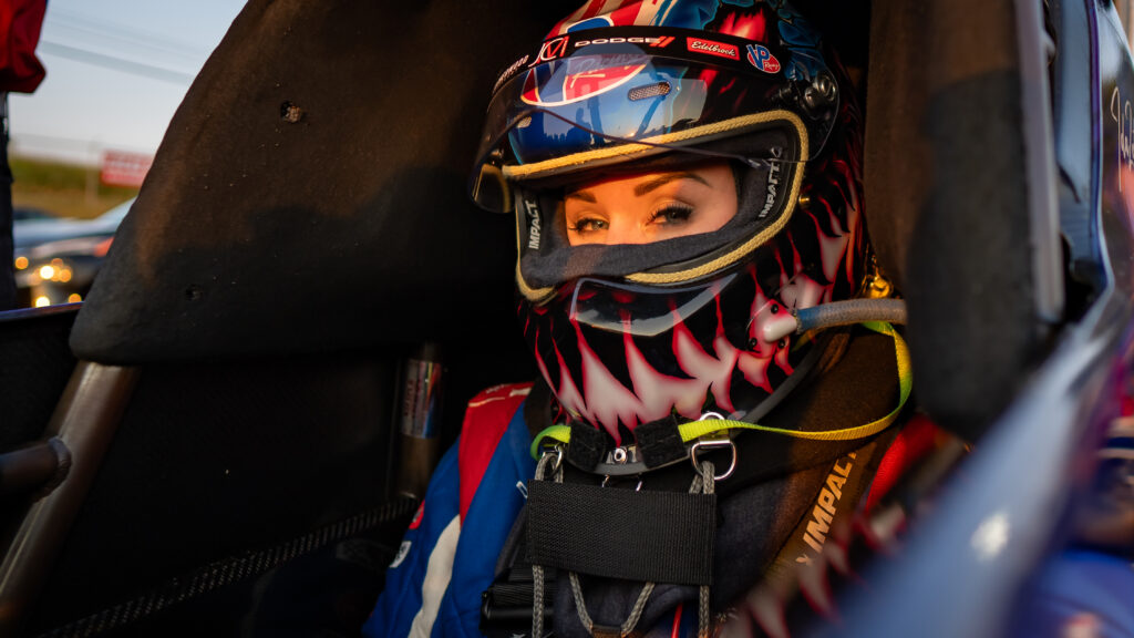 En racerförare med en färgglad hjälm med flammotiv sitter i förarhytten och tittar mot kameran, ordentligt fastspänd i säkerhetsbältena.