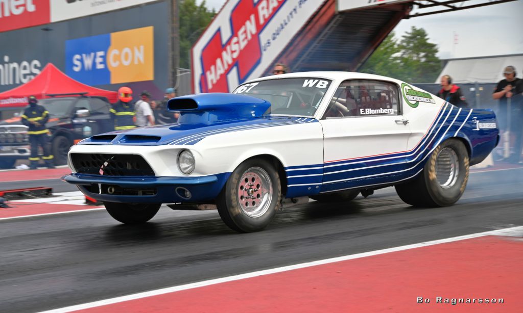 En klassisk blå och vit muskelbil med tävlingsränder som gör en burnout på en dragracingbana, med rök från bakdäcken. I bakgrunden syns säkerhetspersonal och ett rött tält.