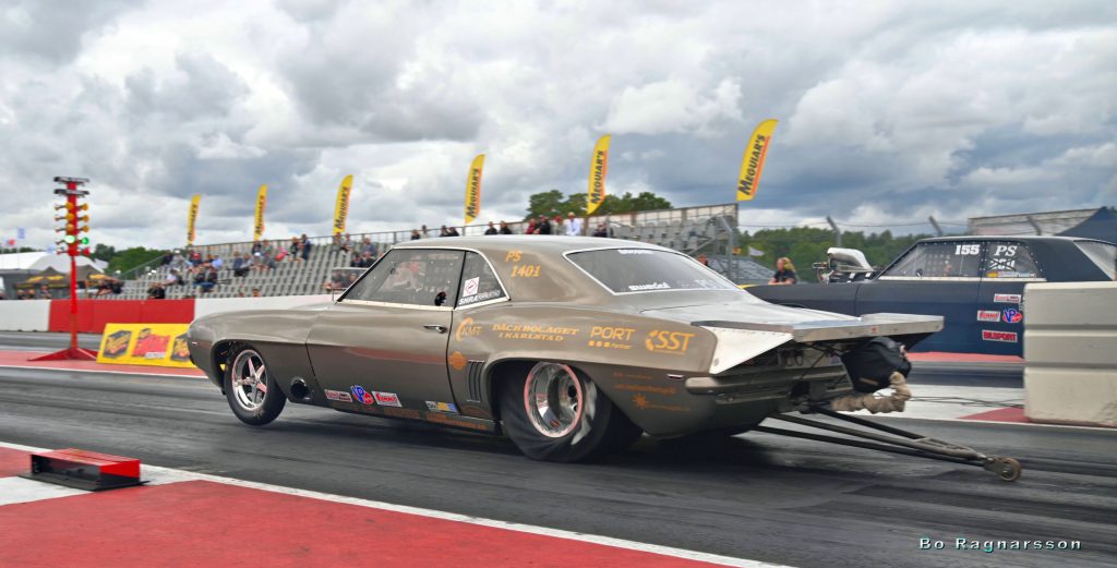 En klassisk dragracingbil accelererar på en racerbana, med bakhjulen lyfta och fallskärmspaketet synligt. Åskådare tittar på från läktarna under en molnig himmel.