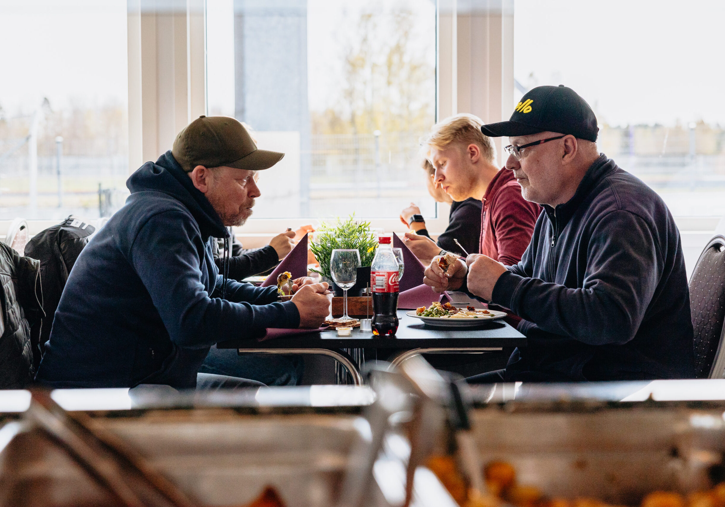 Fyra män sitter vid ett restaurangbord och äter en måltid tillsammans. Tallrikar med mat, drycker och en krukväxt står på bordet. Stora fönster i bakgrunden släpper in dagsljus. Bilden är inramad av brickor med mat i förgrunden.
