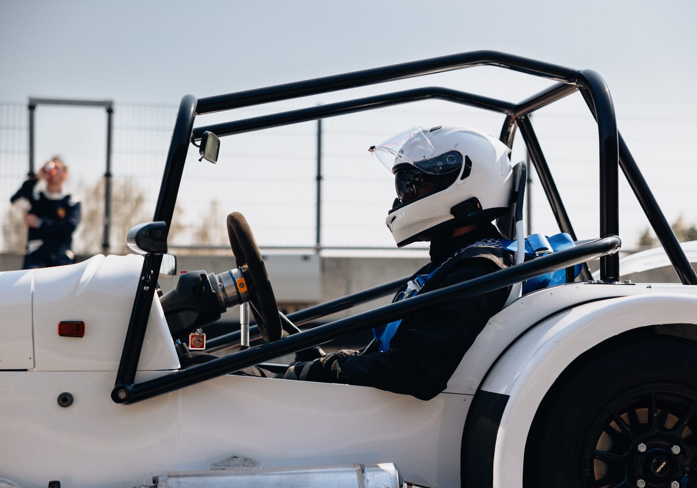 En person med hjälm och tävlingsdräkt sitter i en vit öppen racerbil, redo att köra på en bana. I bakgrunden syns ett staket och en annan person som står på avstånd.