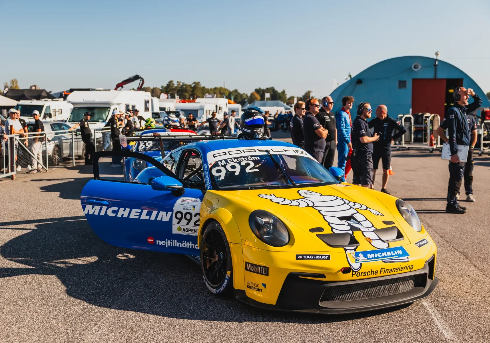 En gul och blå Porsche racerbil med nummer 992 står parkerad vid ett evenemang. Bilen har Michelin-märke. Människor i bakgrunden står nära släpvagnar och en välvd byggnad under en klar himmel.