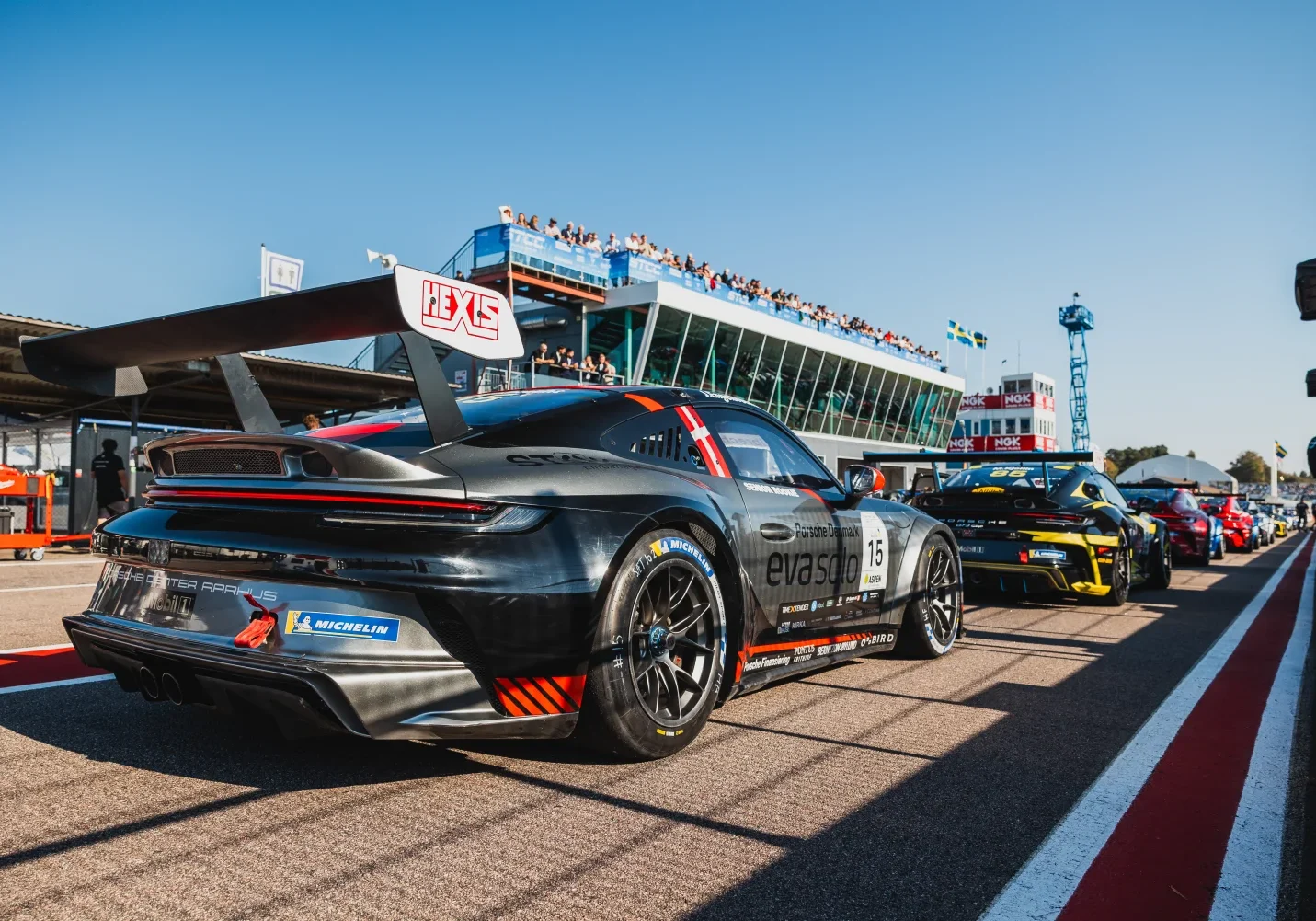 Svart racerbil med stor bakspoiler parkerad på en racerbana. Andra bilar ställde upp bakom. En byggnad med åskådare och flaggor i bakgrunden under en klarblå himmel.