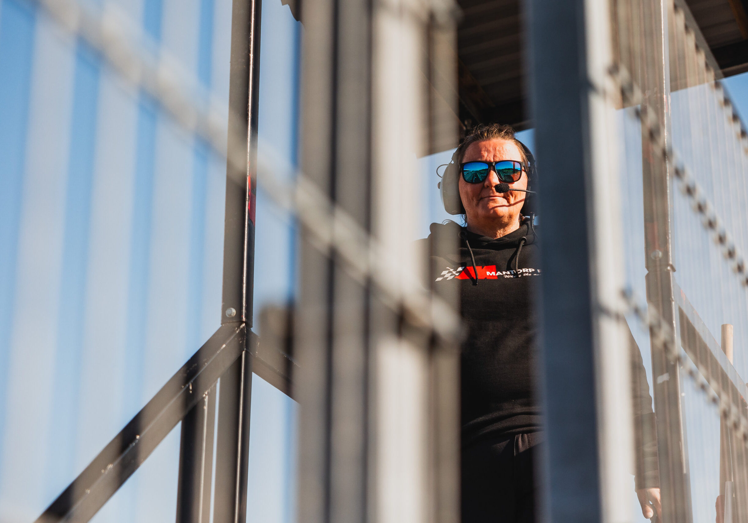 En person med solglasögon och svart huvtröja står på en metalltrappa, delvis skymd av metallstänger, med blå himmel i bakgrunden.