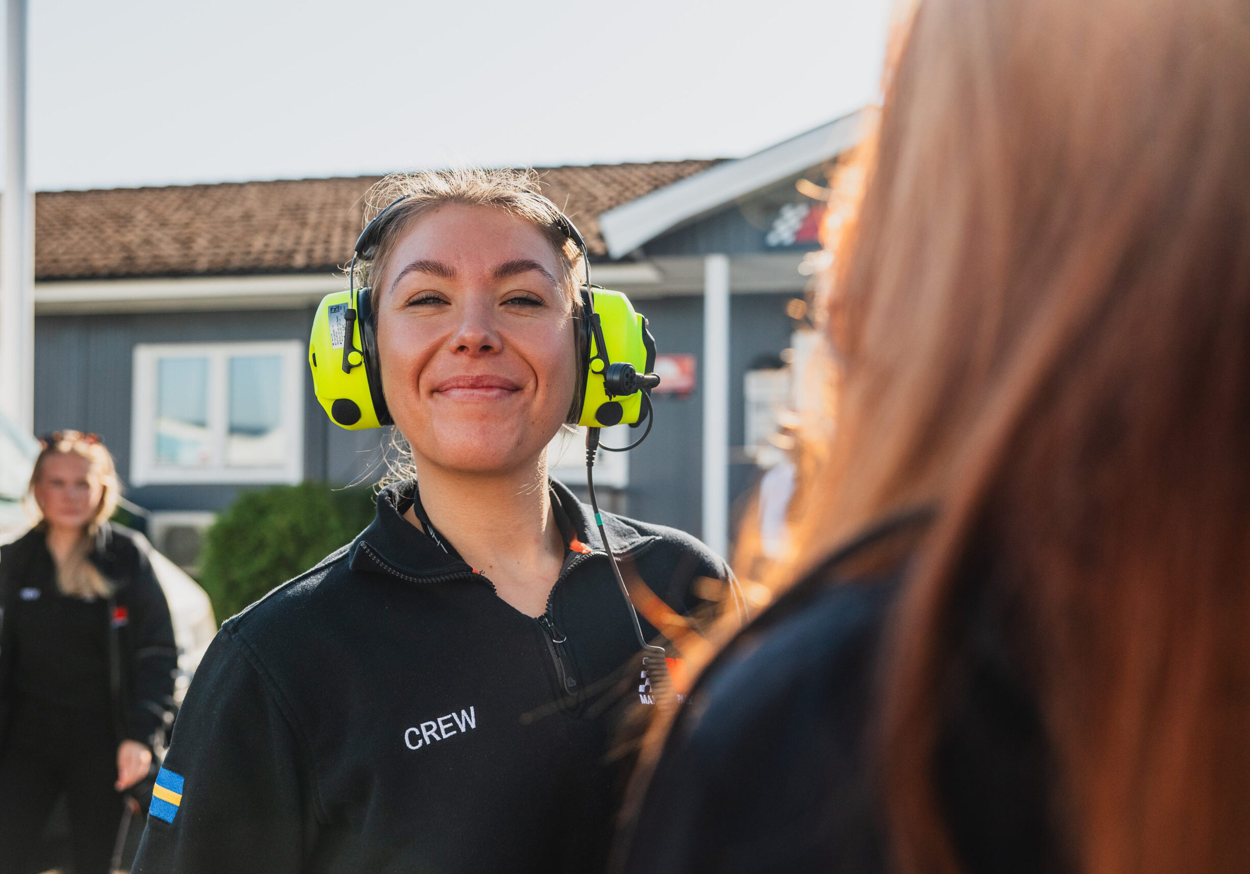 En kvinna med headset och jacka med CREW-märkning ler mot kameran utomhus, med en annan person i förgrunden och en byggnad i bakgrunden en solig dag.