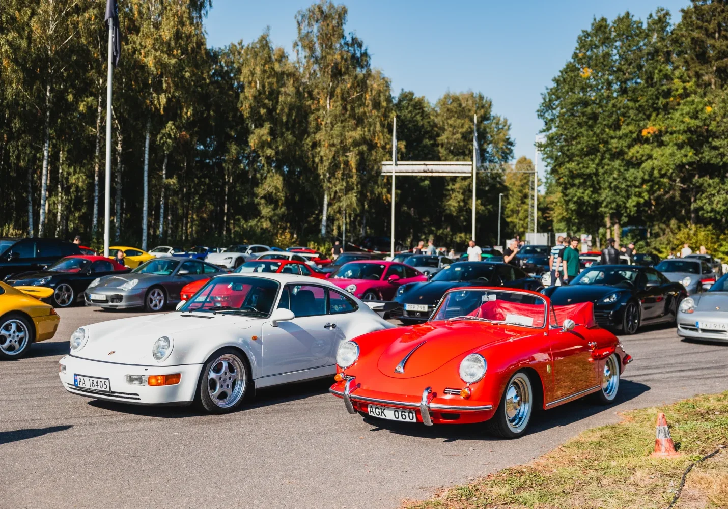 En samling klassiska bilar på en parkeringsplats med en vit coupé och en röd cabriolet framtill. Flera andra färgglada bilar står parkerade i bakgrunden, omgivna av träd och människor en solig dag.