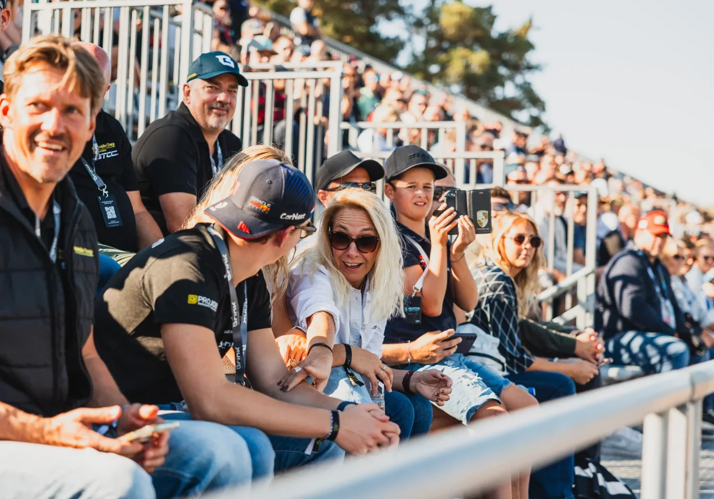En grupp människor sitter på läktaren vid ett utomhusevenemang. De ler och pratar, med några som bär hattar och solglasögon. I bakgrunden tittar fler deltagare från läktaren under klar himmel.
