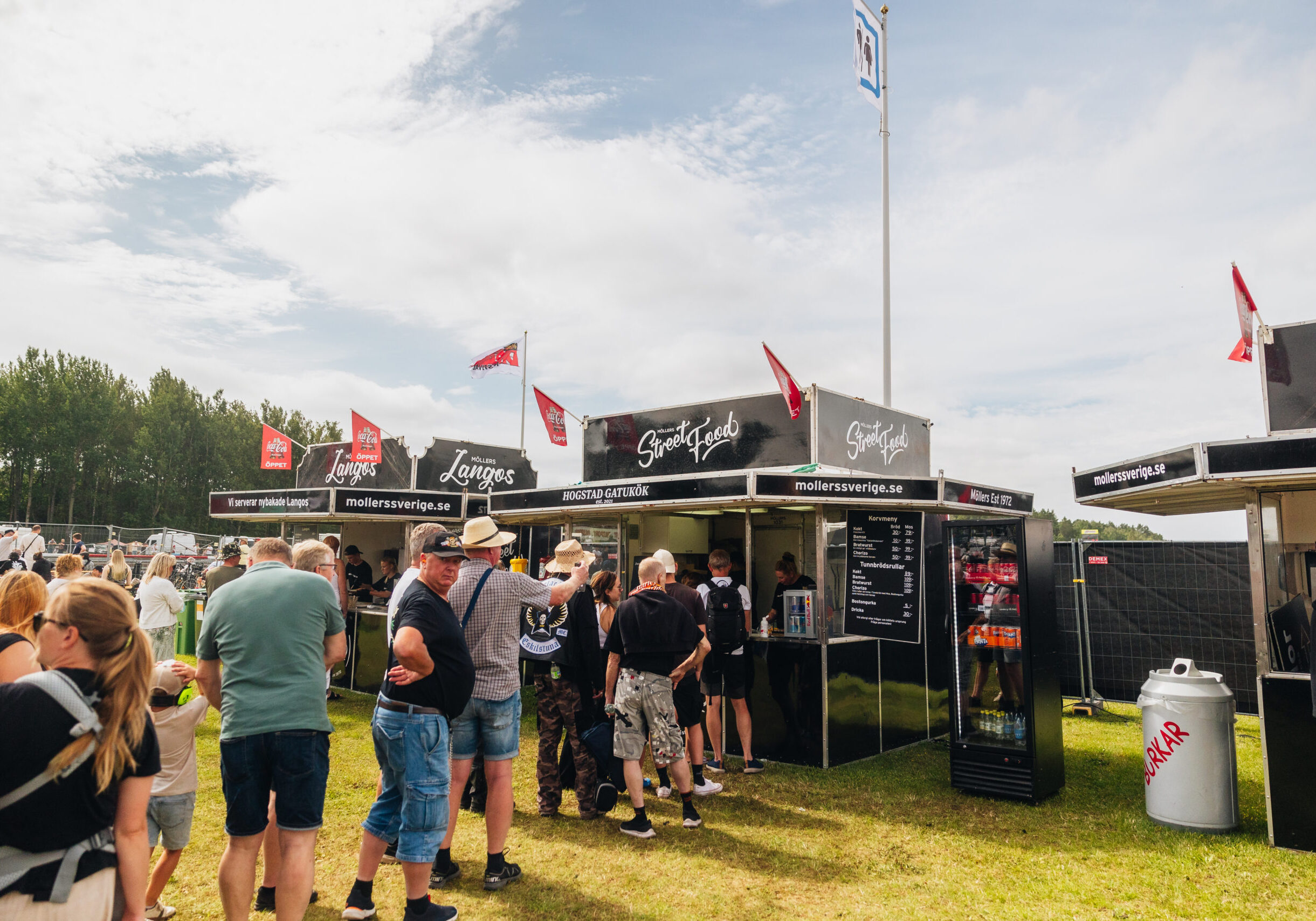 Människor står i kö vid ett utomhusmatstånd med svarta skyltar och röda flaggor på en gräsplan under en delvis molnig himmel, med träd och andra festivalbesökare i bakgrunden.