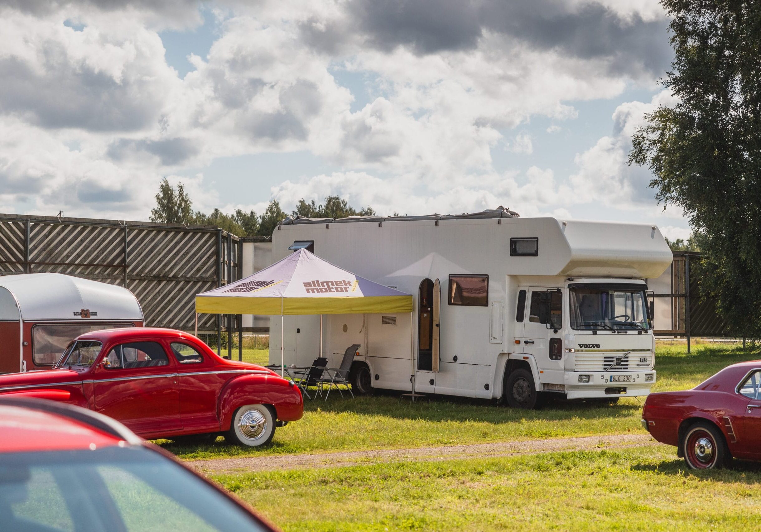 En stor vit husbil står parkerad på gräset bredvid ett tält med skärmtak. Röda veteranbilar står parkerade i närheten, med träd och en molnig himmel i bakgrunden.
