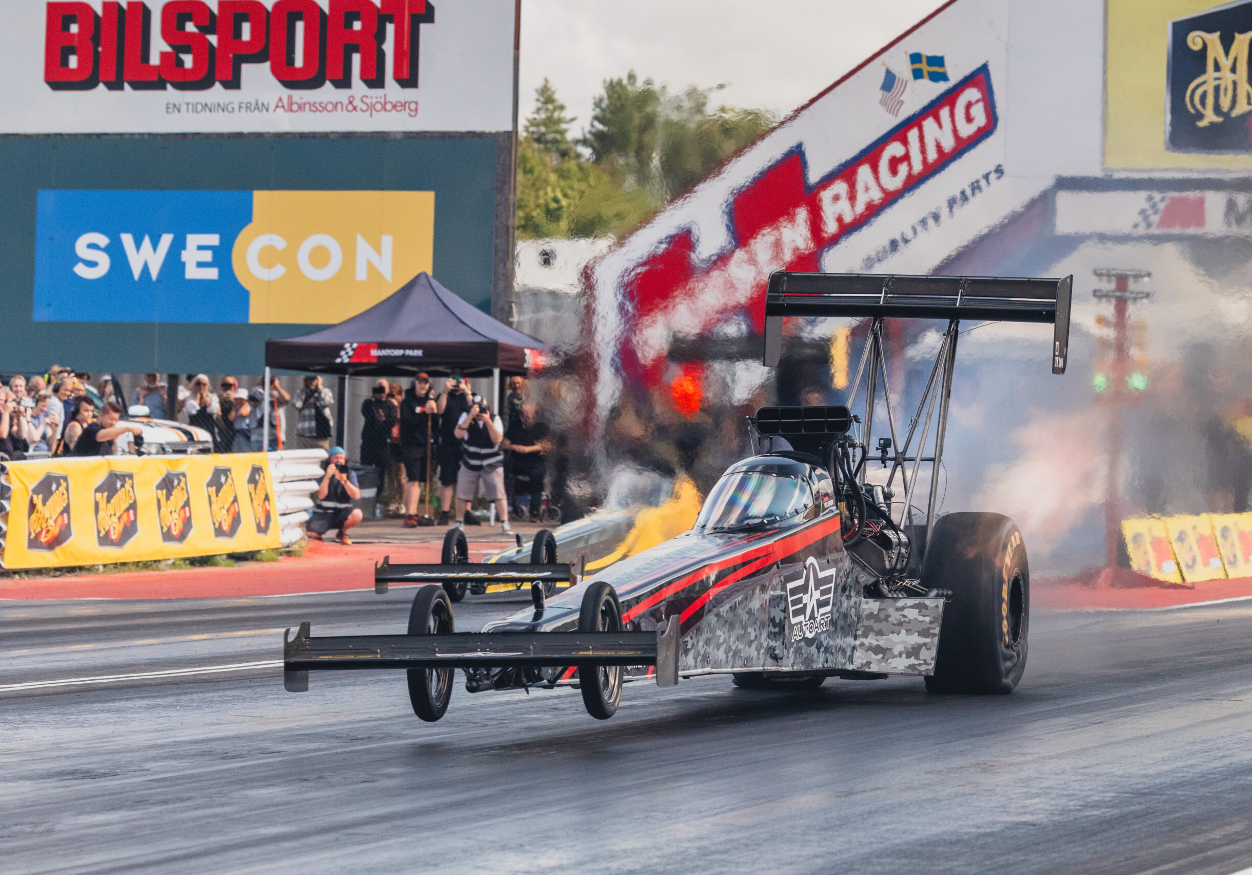 En dragracingbil accelererar nedför en bana med rök från däcken, med en publik och sponsorbanderoller i bakgrunden.