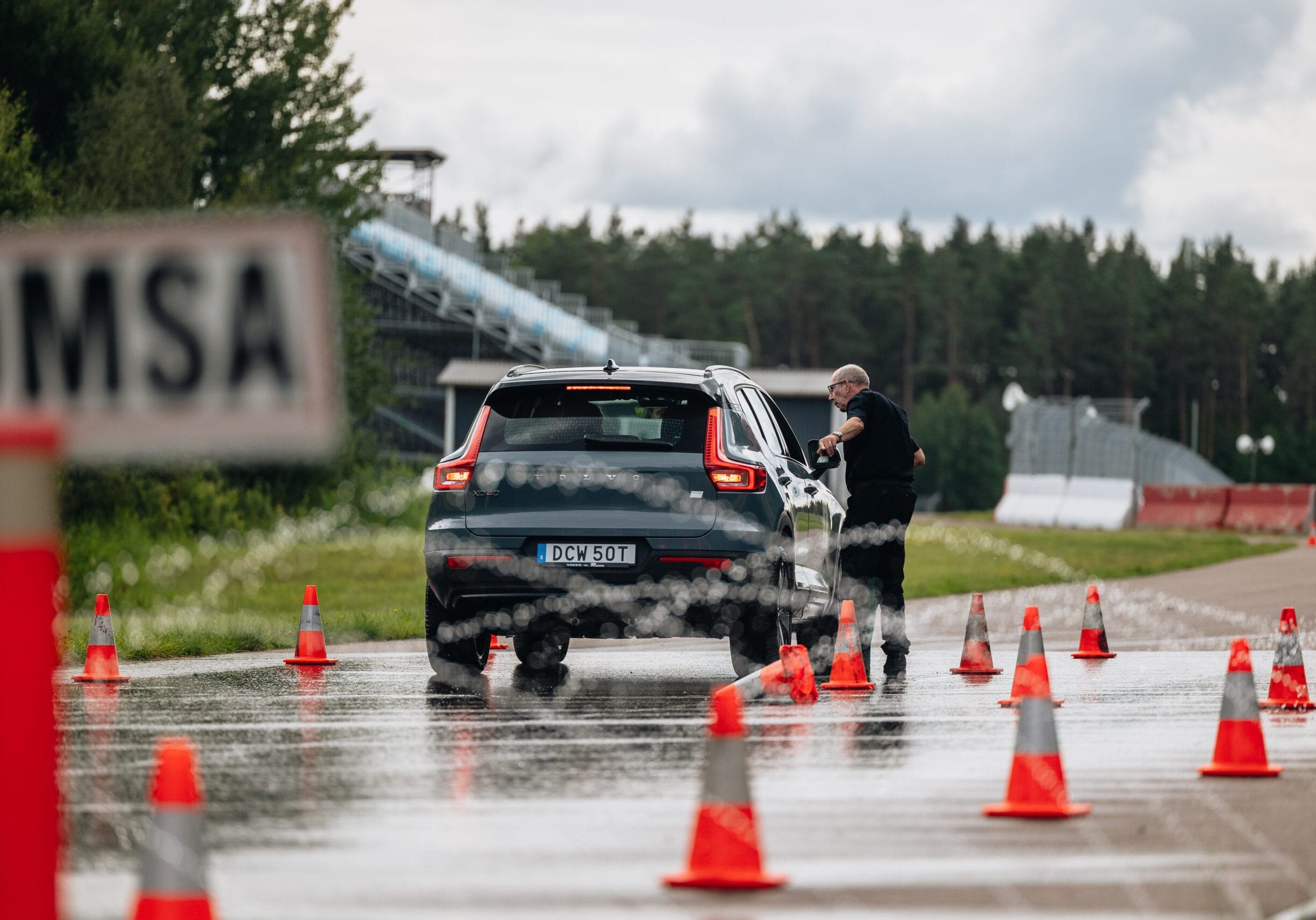 En person står bredvid en mörk SUV på en våt väg kantad av orange trafikkoner, troligen vid ett körprov eller en utbildning, med en skog och molnig himmel i bakgrunden.