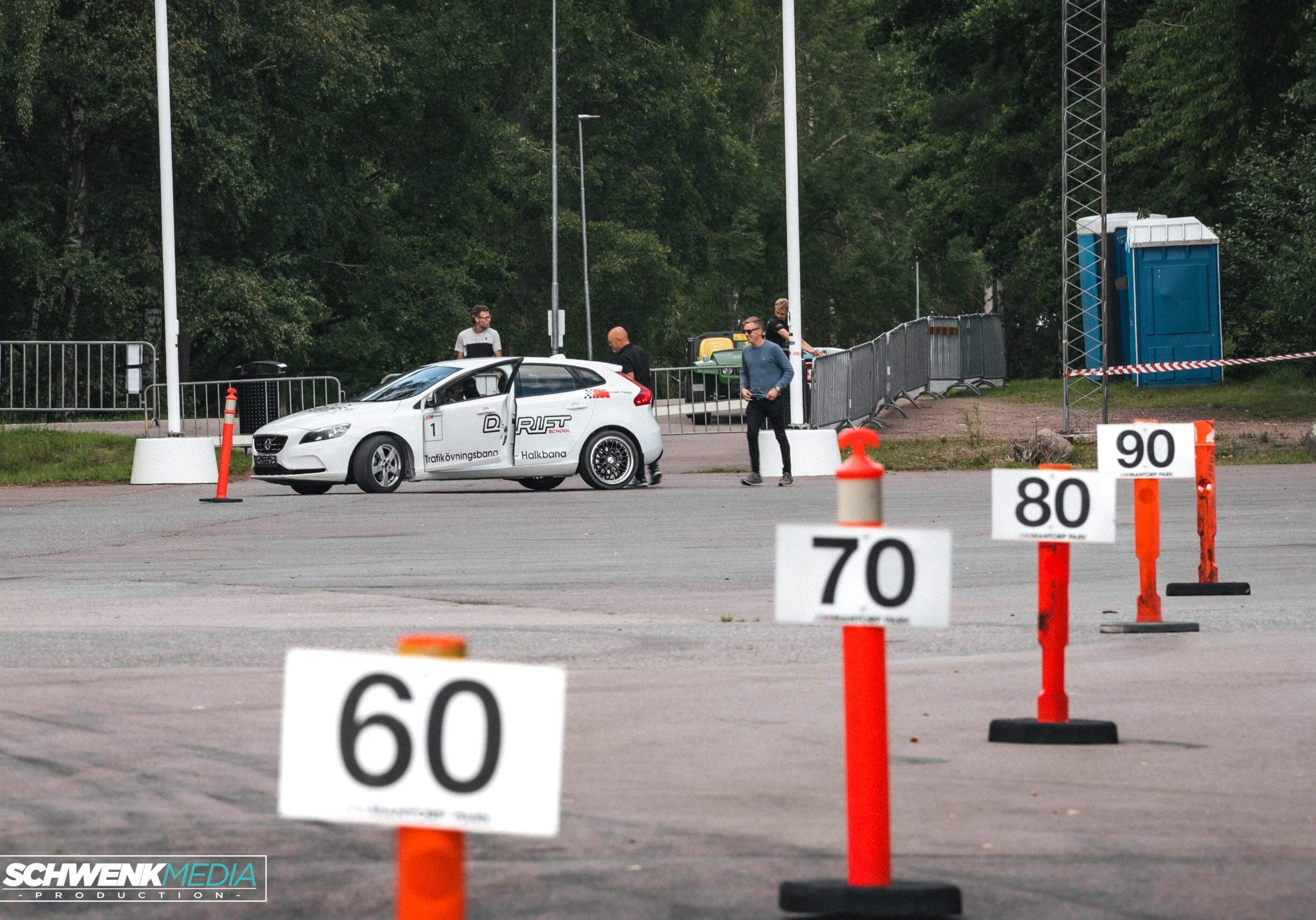 En vit bil med B Riet-märkning är parkerad nära två personer som står framför den. Orangea koner med skyltar för hastighetsbegränsning (60, 70, 80, 90) står uppradade på asfalten i förgrunden. Träd finns i bakgrunden.