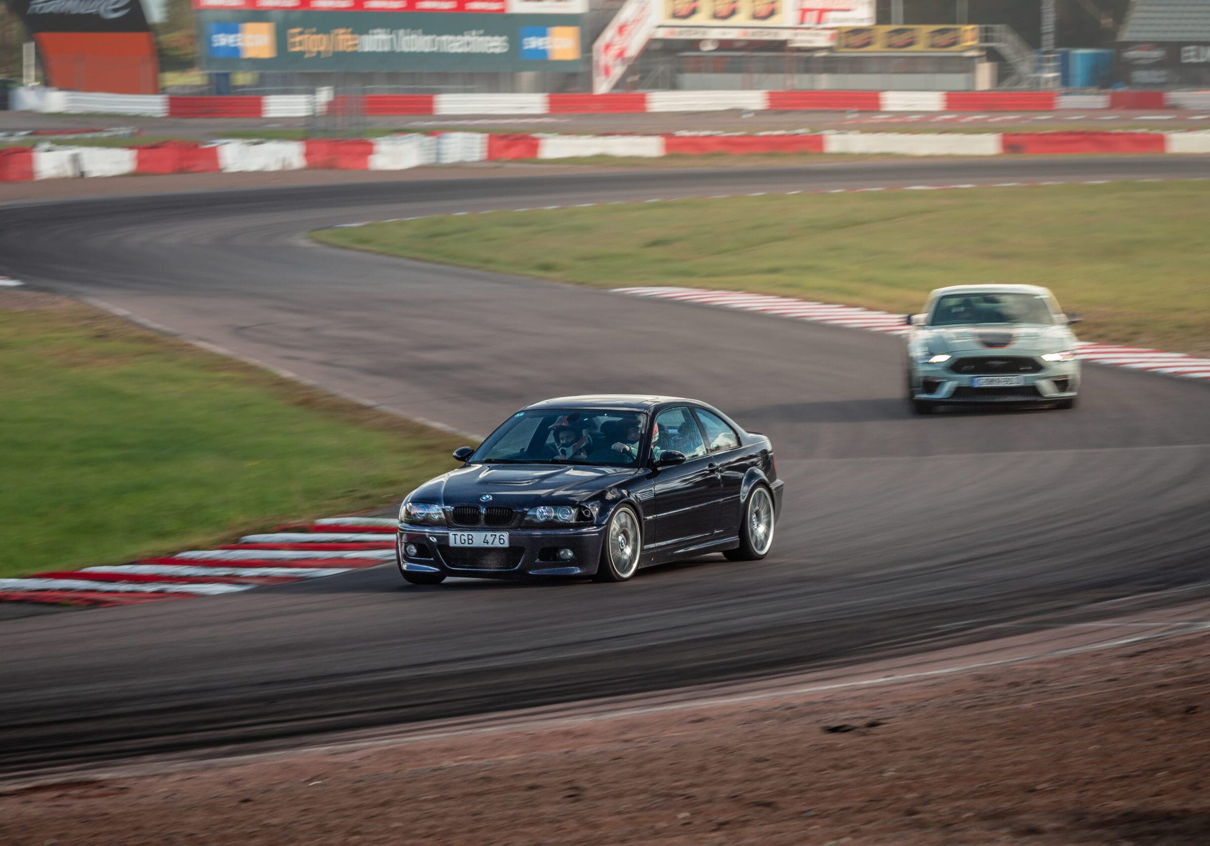 Två bilar tävlar runt en kurva på en racerbana, med en mörkblå bil framför och en vit bil bakom. Banan är omgiven av gräs, bommar och reklamskyltar i bakgrunden.