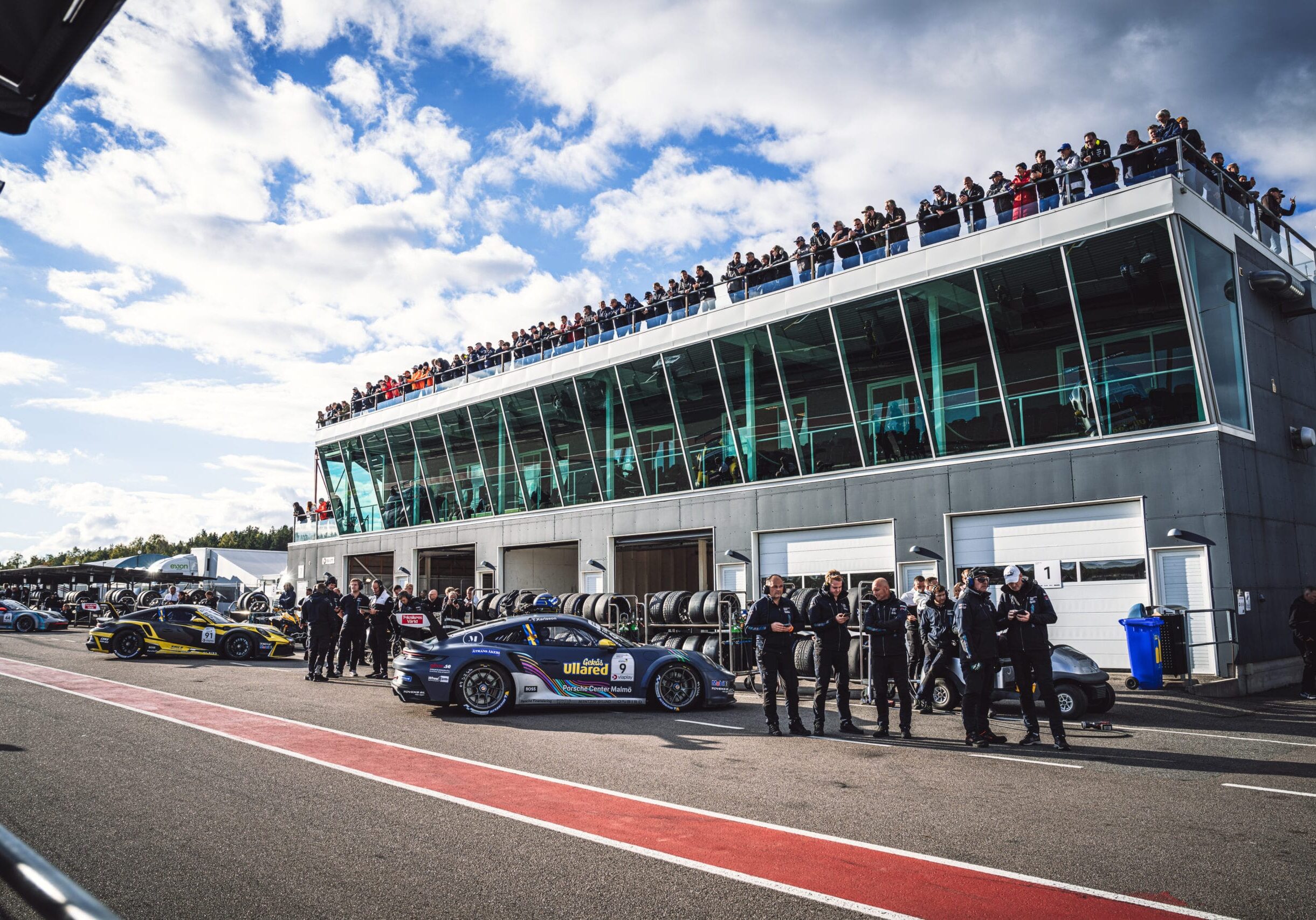 Racerbilar och förare står uppradade på en bana nära en modern glasbyggnad med stora fönster. Åskådare tittar på från taket och himlen är delvis molnig. Tävlingsdäck och garageportar syns i bakgrunden.