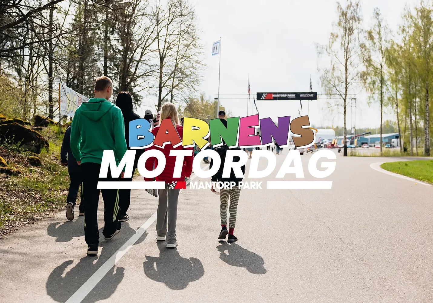 En grupp människor, inklusive barn, går längs en asfalterad stig mot en startlinje. Orden "Barnens Motordag Mantorp Park" står väl synligt över bilden. Glesa träd och grönska syns längs stigen.