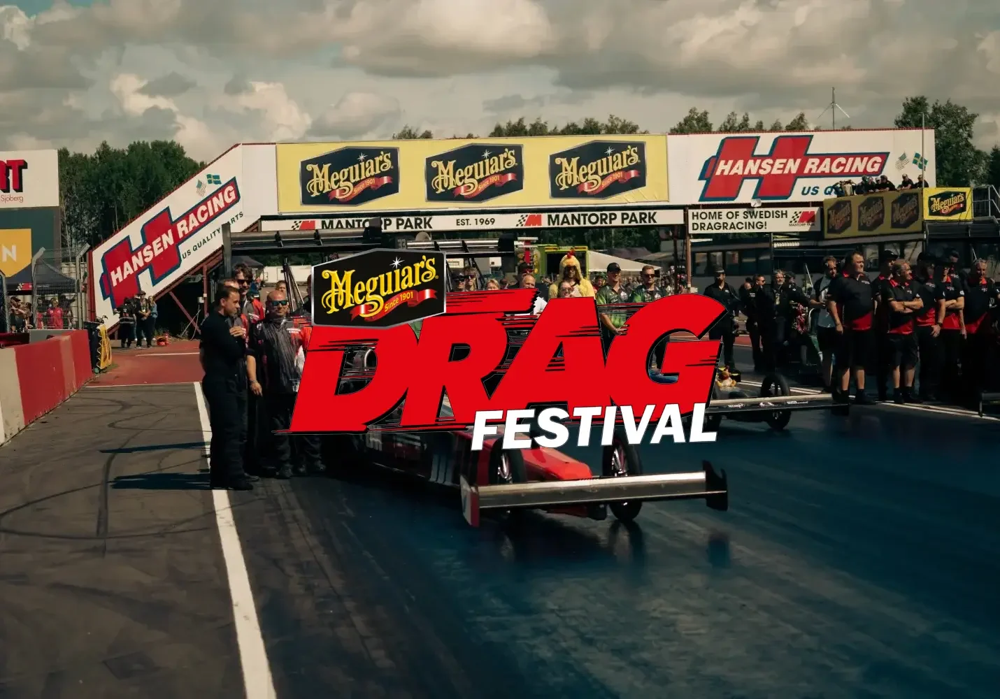 Dragracing-scen på Mantorp Park med en dragster redo att tävla. Stor publik och personal på varje sida. Meguiar's och Hansen Racing banderoller visades framträdande. "DRAG Festival"-text överlagrar bilden. Molnig himmel i bakgrunden.