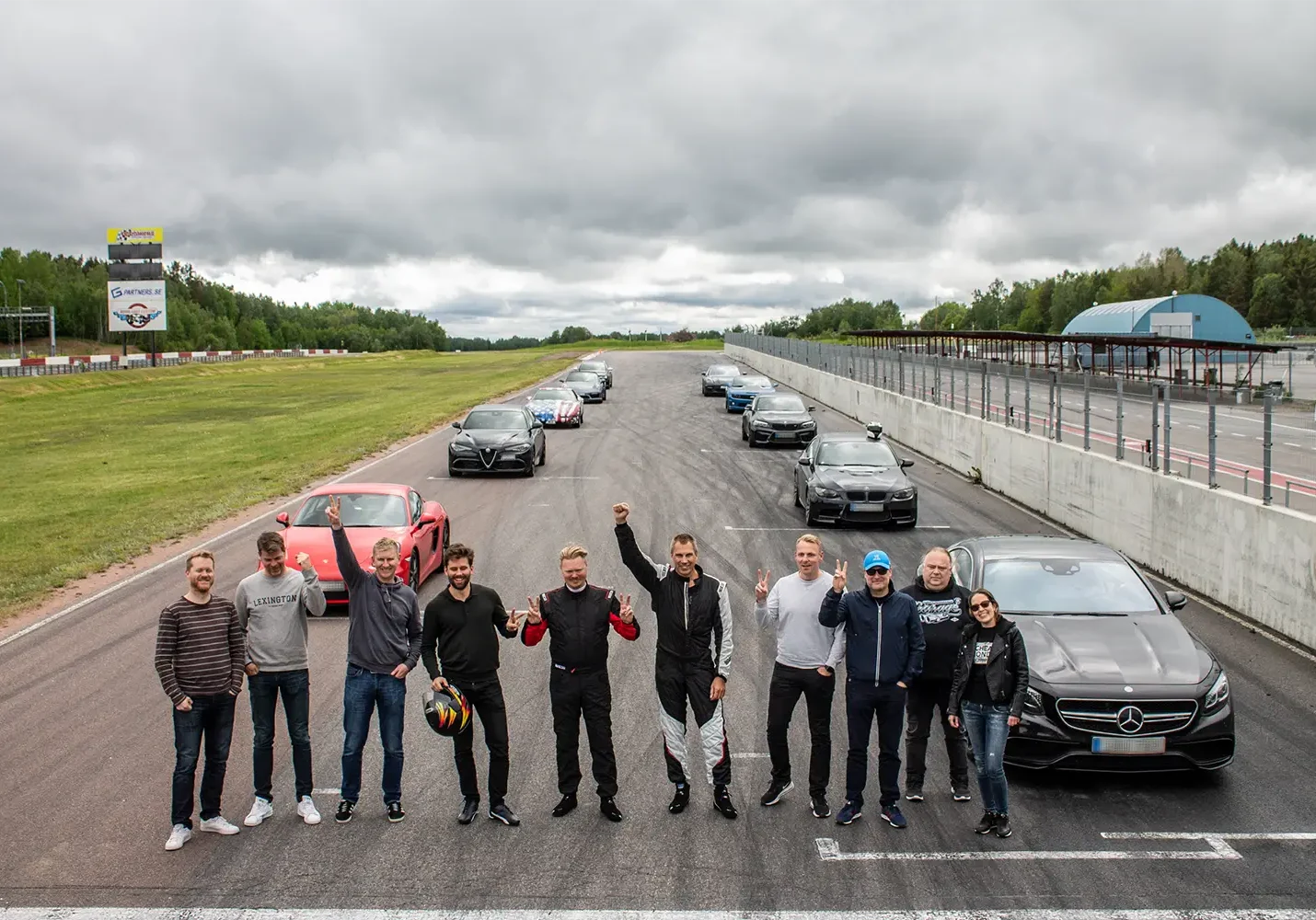 En grupp människor, några i racingutrustning, står och ler på en racerbana med en rad sportbilar bakom sig. Himlen är mulen, och det finns gräs och träd till vänster och en byggnad till höger.